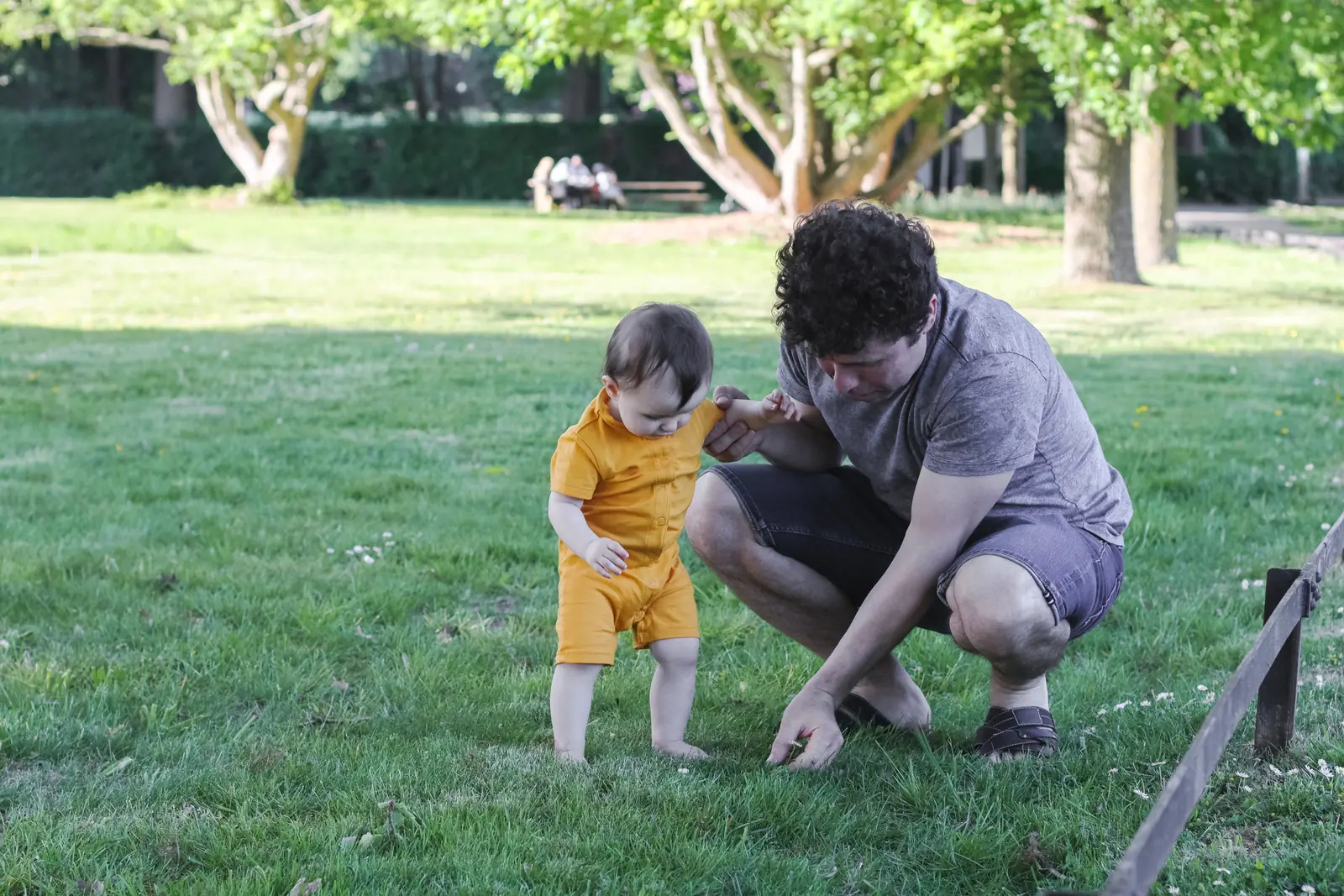 Adult crouching beside a toddler in a yellow outfit as they look at the grass together