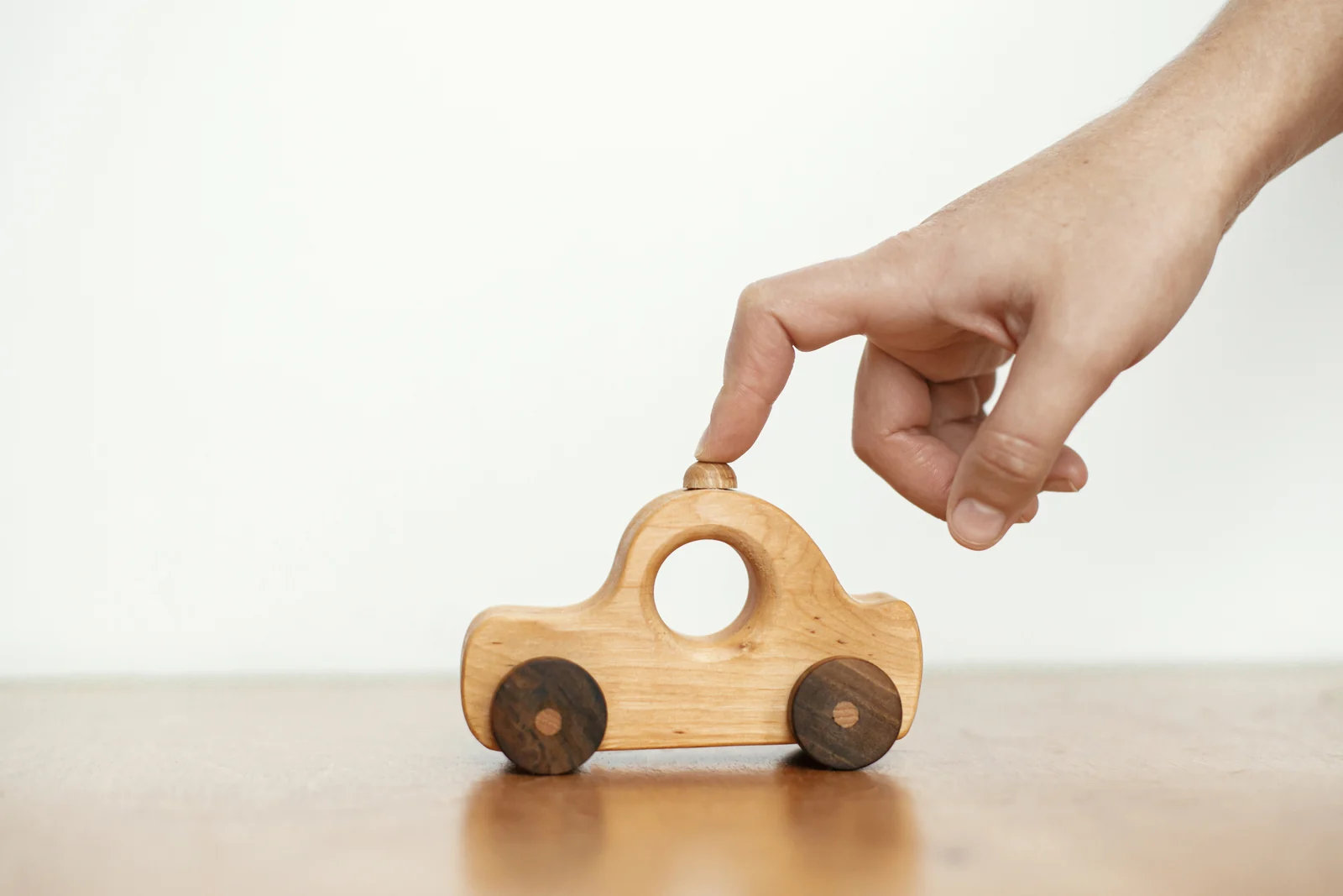 An adult's fingertip rests on a smooth wooden toy car sitting on a table