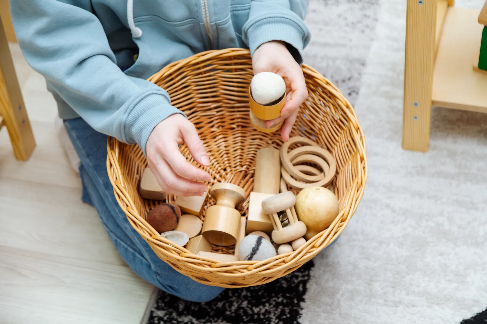 Adult hands sorting plain wooden toys, rings and a wooden rattle in a woven basket on the floor