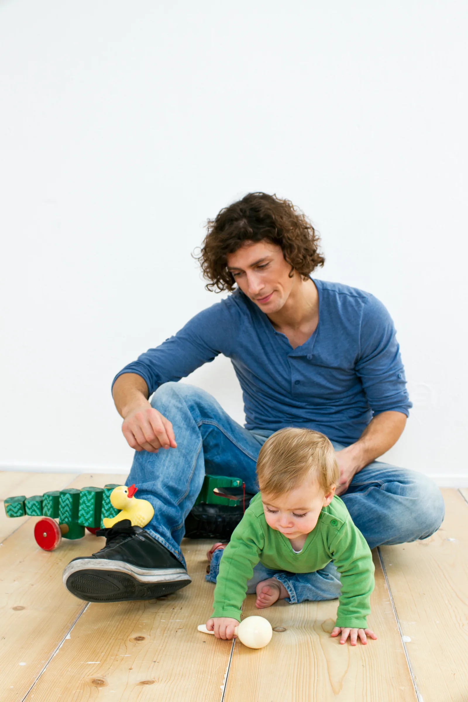 An adult sits on a wooden floor right beside a crawling baby who is reaching for a wooden toy
