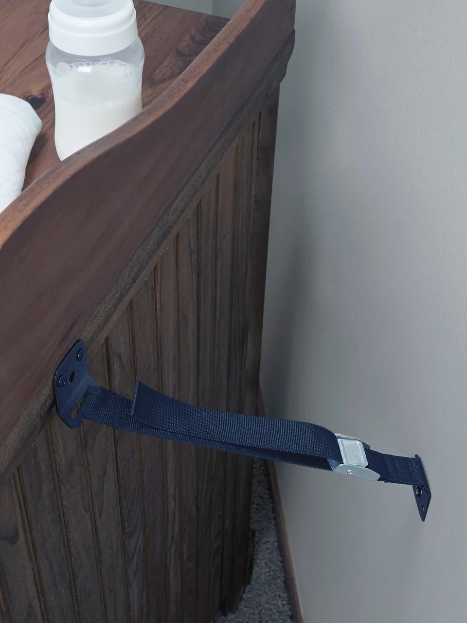 A navy anti-tip strap anchoring a wooden dresser to the wall, with a baby bottle on top