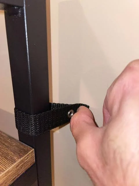 A hand holds a black anti-tip strap looped around the frame of a shelf against a wall