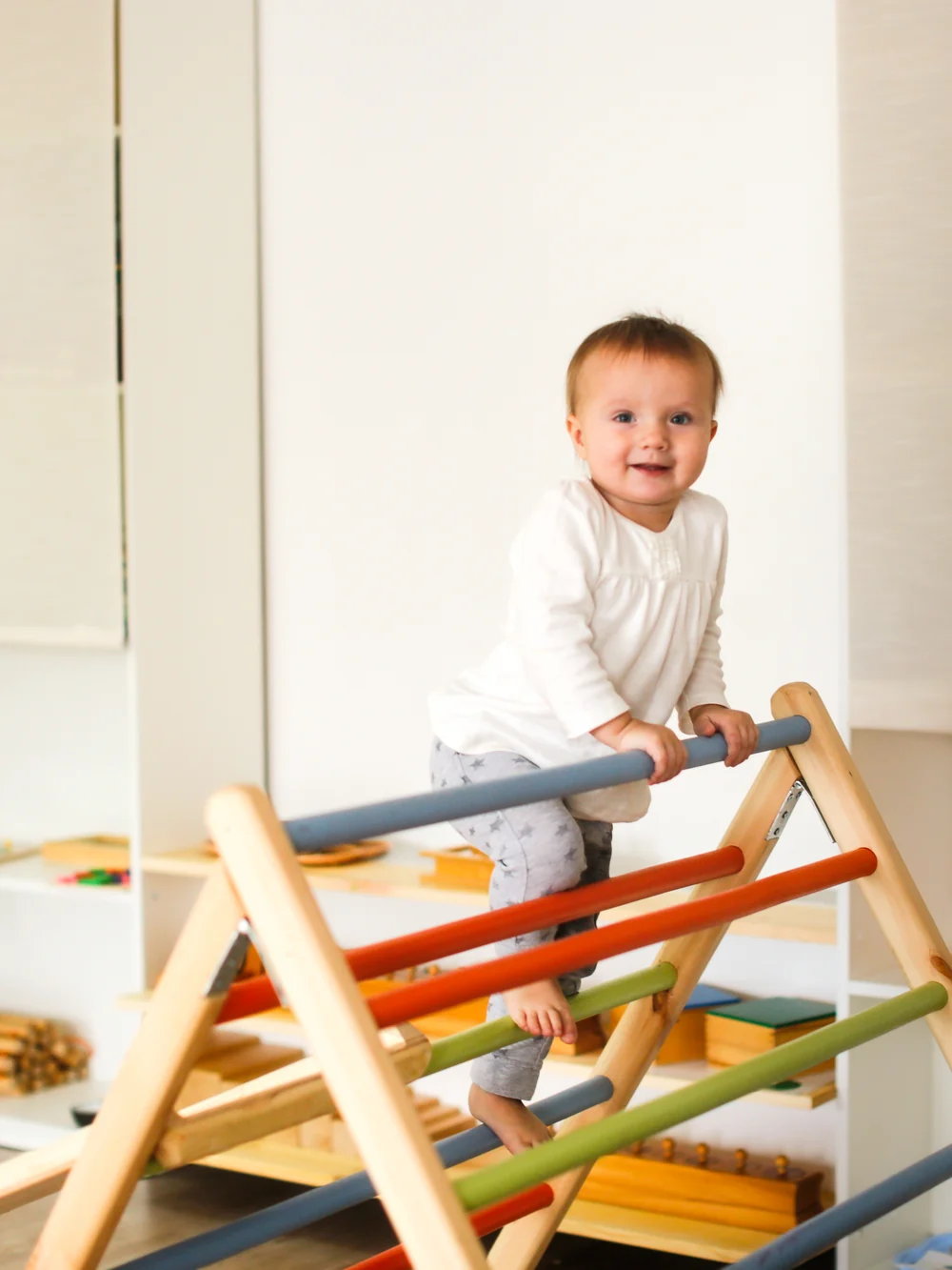 A baby in white stands on the rungs of a colorful wooden Pikler climbing triangle in a living room