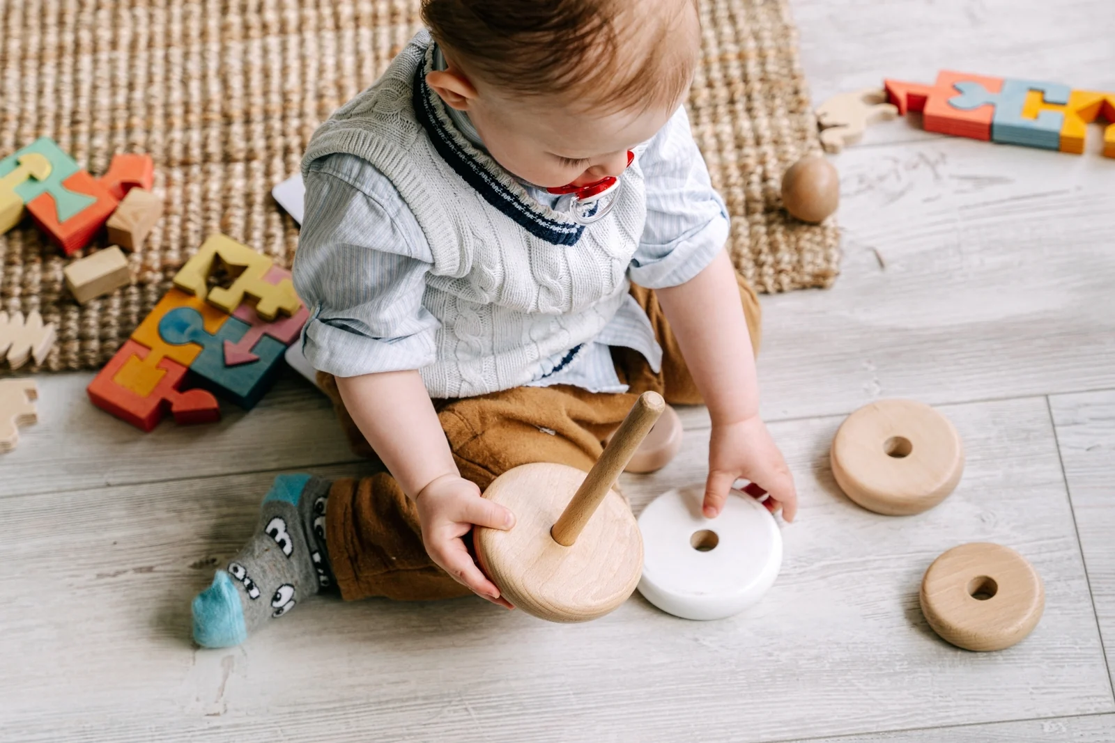 A baby in a knit vest sits on a wood floor holding a wooden ring from a stacking toy