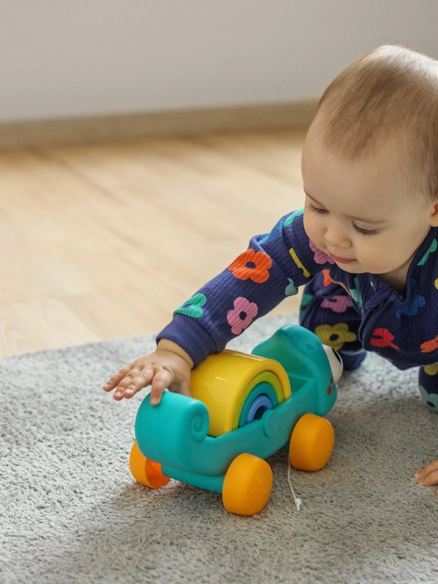 A baby in a floral outfit crawls on a gray rug toward a colorful wheeled toy