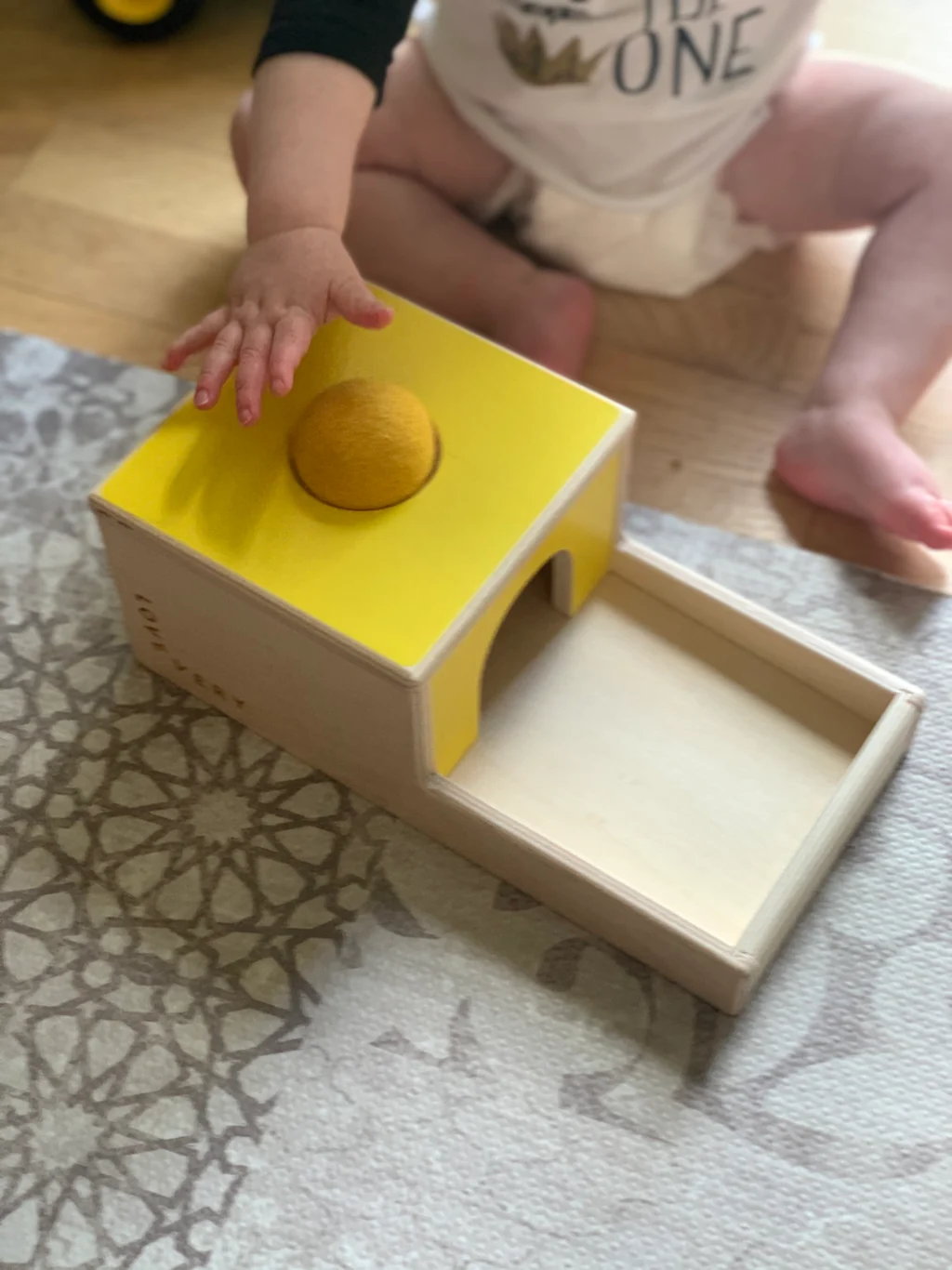 A baby's hand rests on the ball on top of a yellow and wood ball-drop box with an open tray