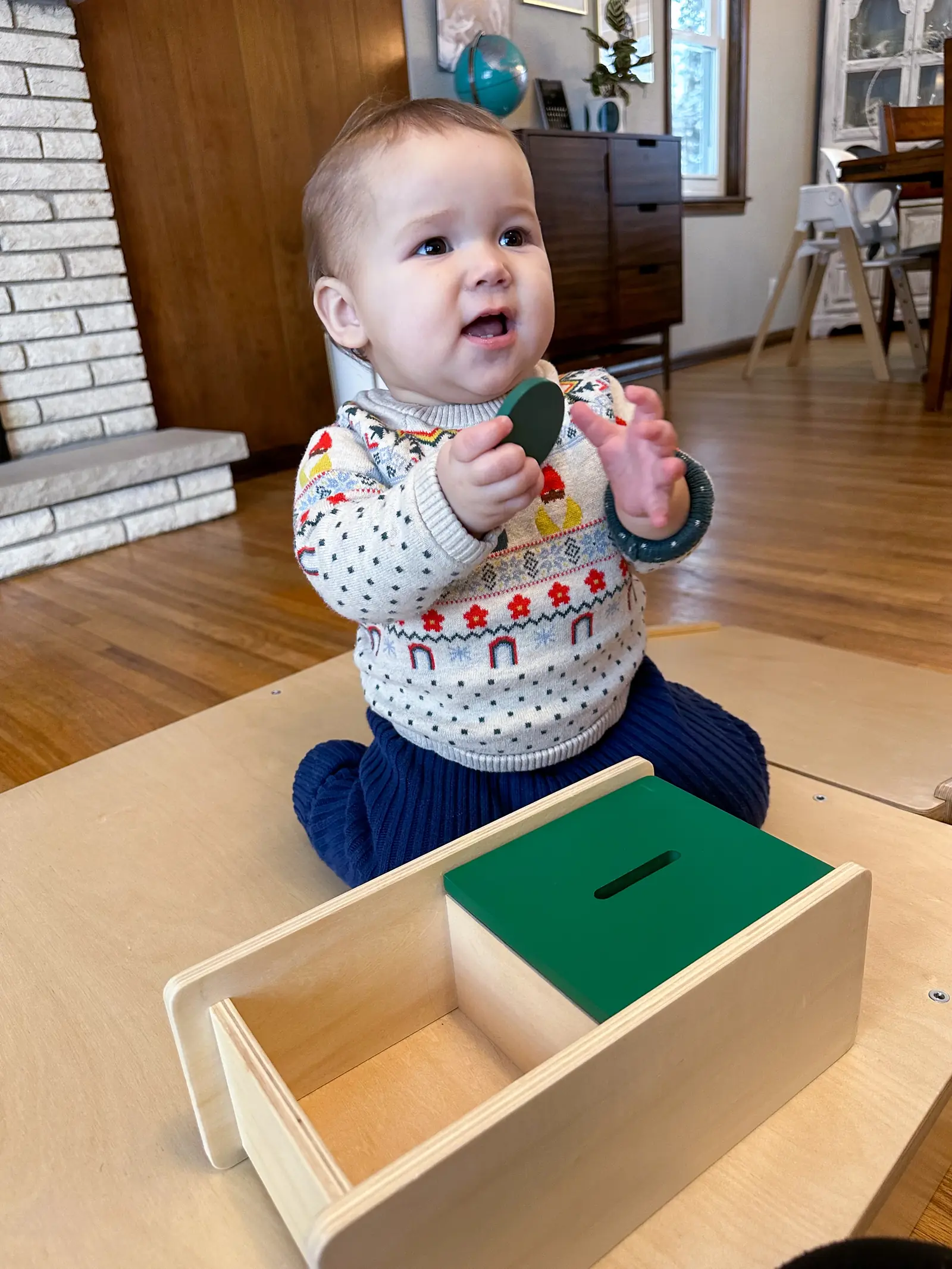 A baby in a patterned sweater sits behind a wooden box with a slotted green lid, holding up a flat green disc