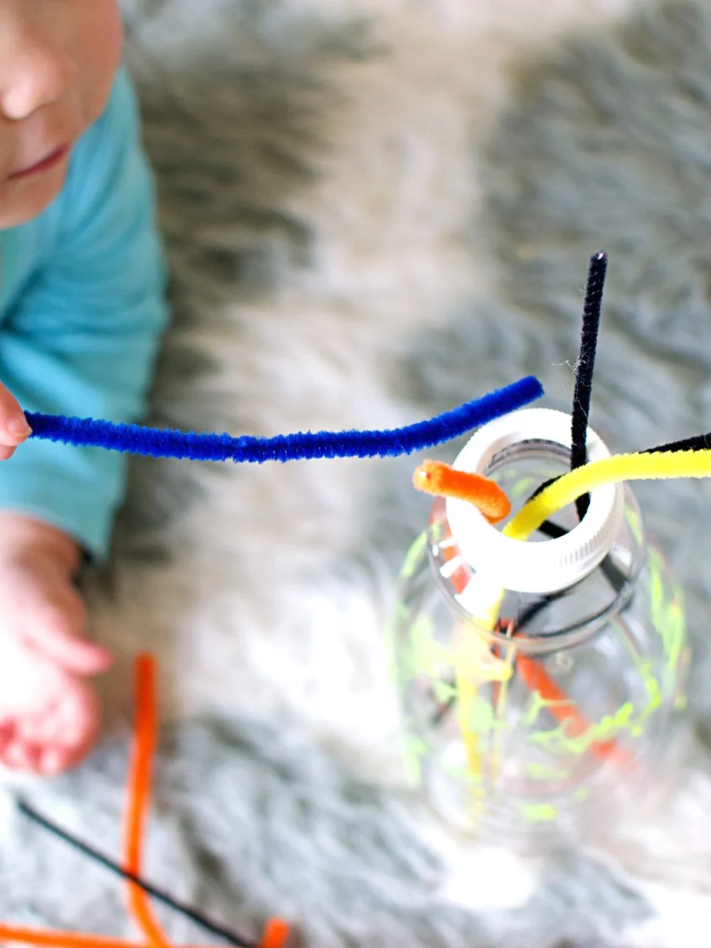 Baby on a fluffy rug with a blue pipe cleaner in hand, next to a clear bottle with pipe cleaners poking out of the top