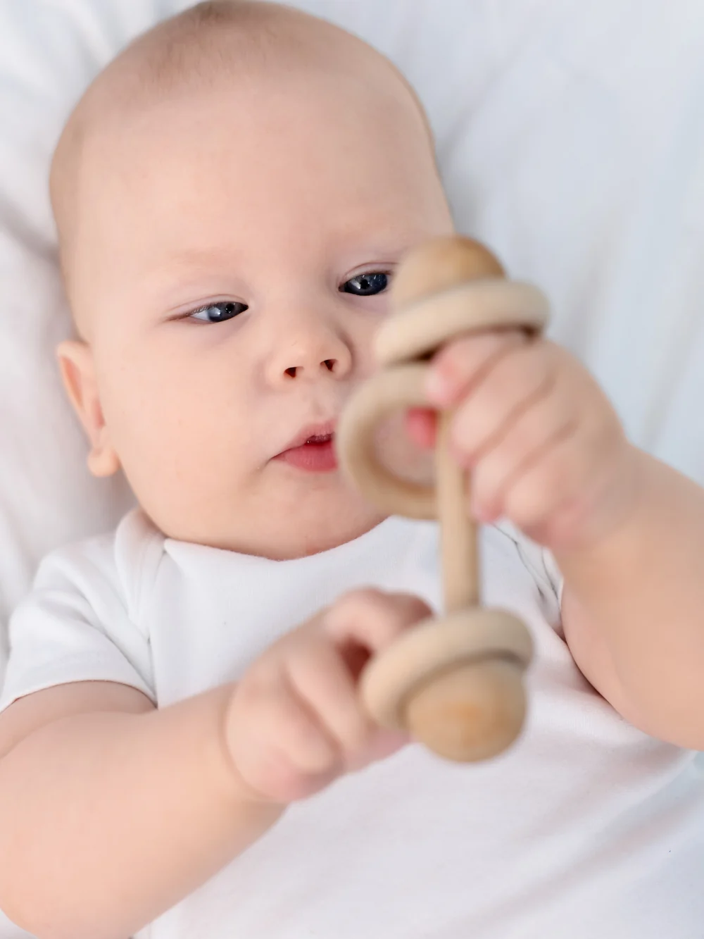 Baby lying on a white blanket holding a wooden ring rattle with both hands