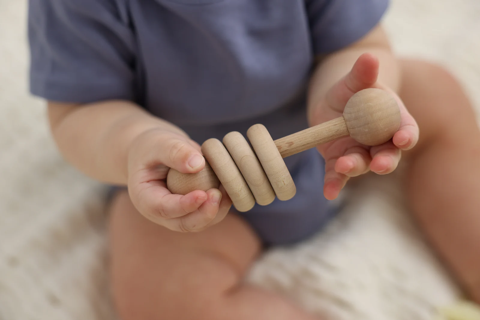 Baby's hands holding a wooden rattle with three smooth rings, seated on a cream blanket
