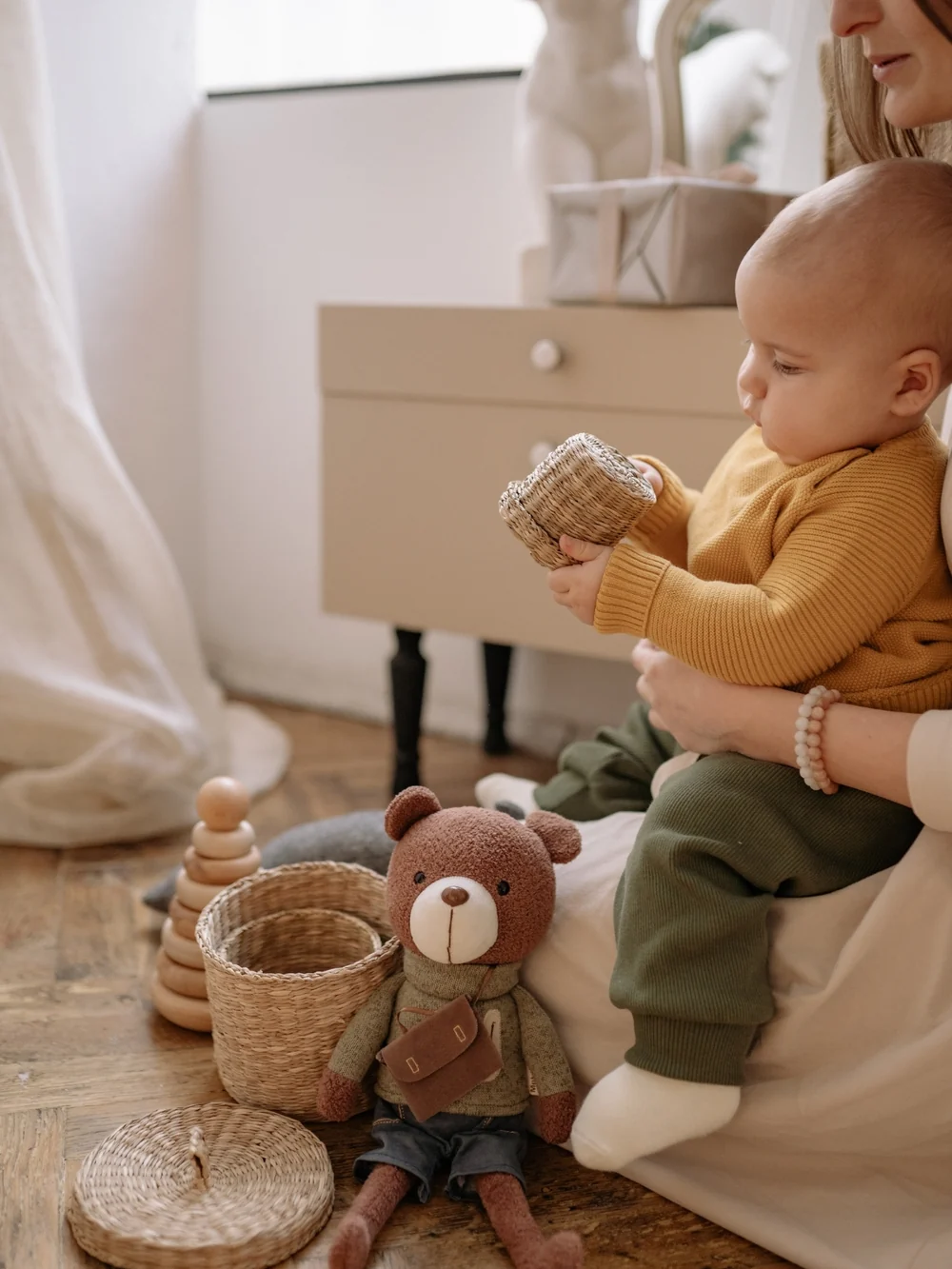Baby on a parent's lap holding a small woven basket, with wooden stacking rings and a teddy bear on the floor