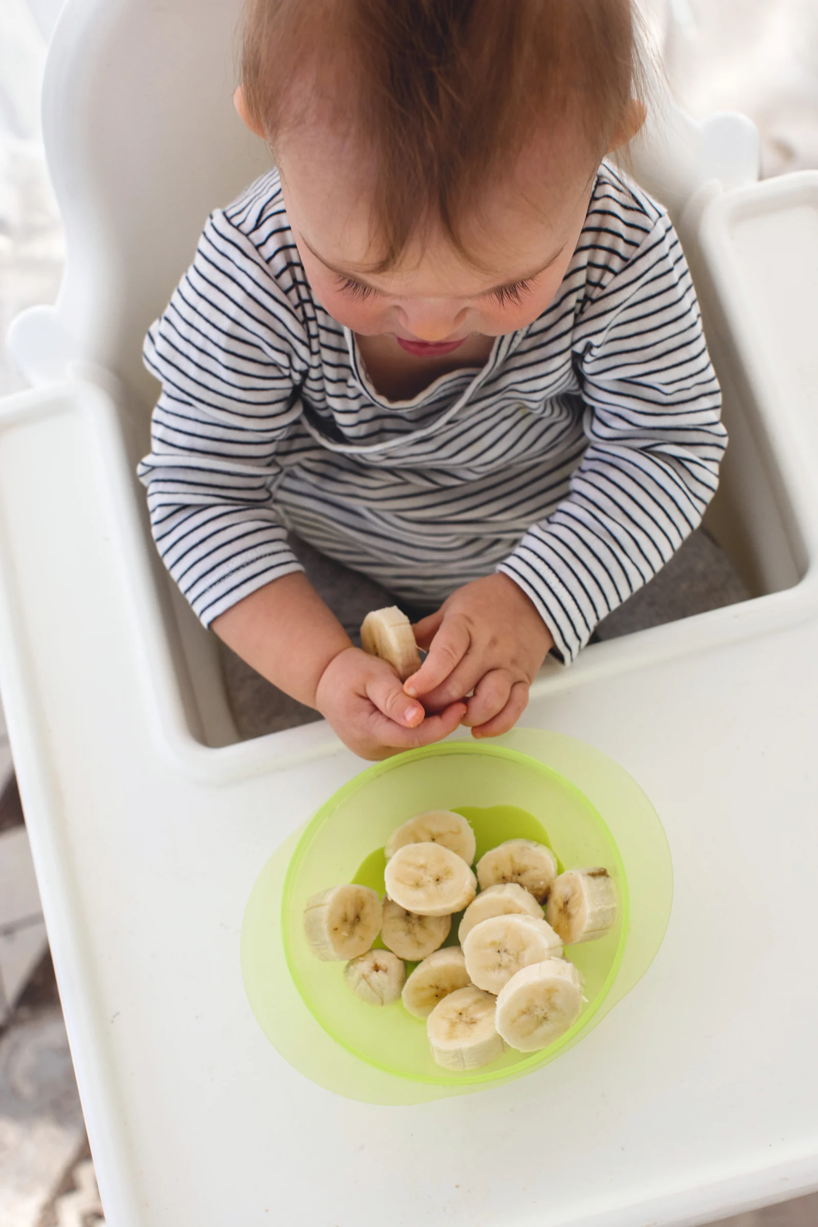 Baby in a white high chair holding a banana slice beside a green bowl of sliced banana