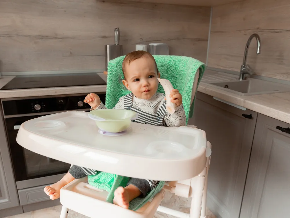 A baby in a high chair holds a piece of banana, with a bowl on the tray