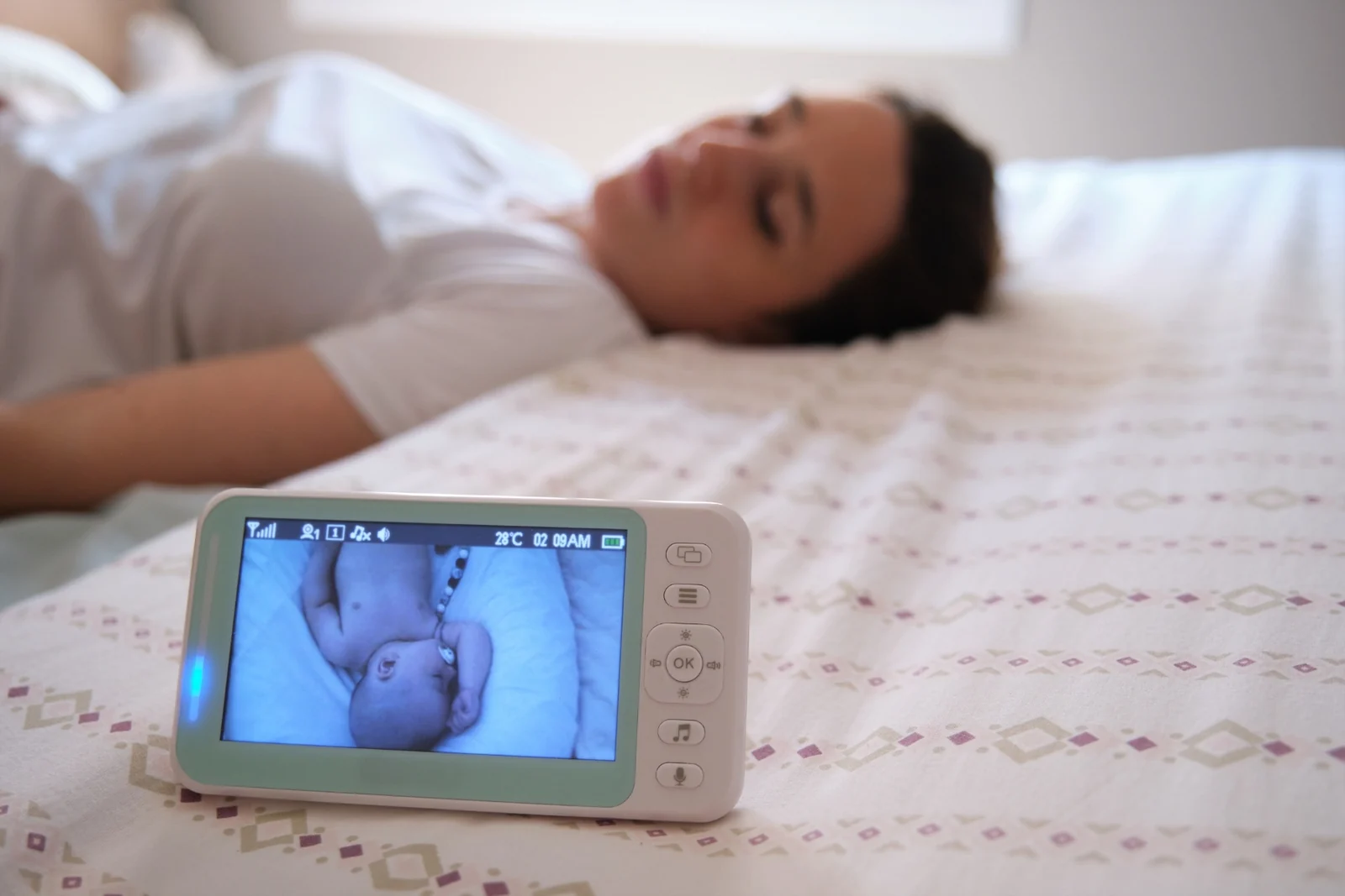 A baby monitor showing a baby on its screen, set on a bed beside a woman lying with her eyes closed