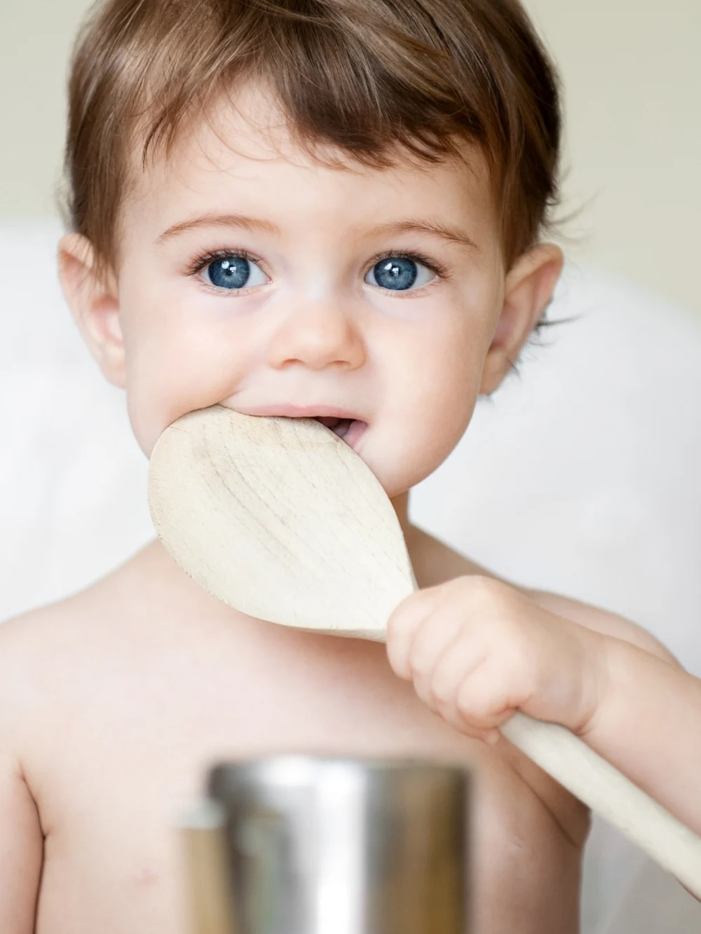 A baby chews on the end of a large wooden spoon, with a steel cup blurred in front