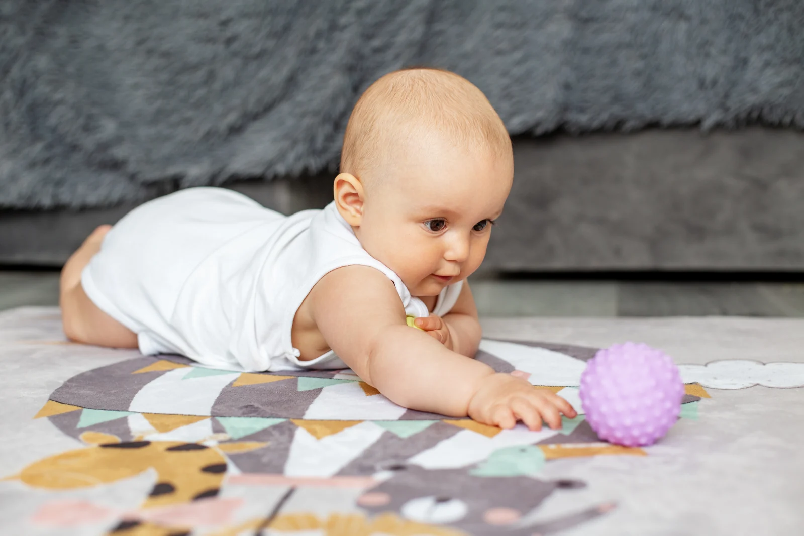 Baby lying on a play mat on the floor, reaching toward a single wooden toy