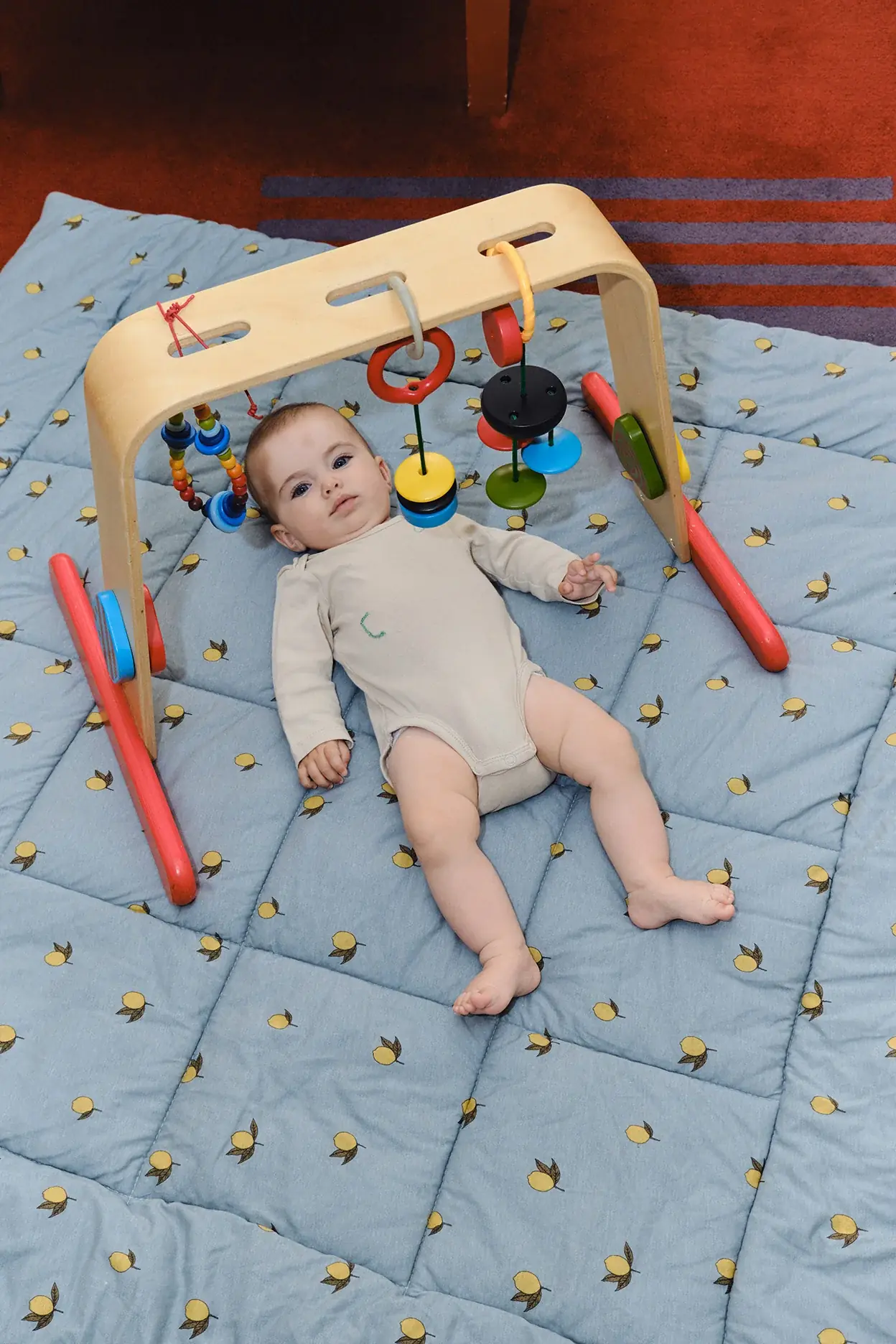 A baby in a cream bodysuit lies on a quilted play mat under a wooden play gym with hanging toys