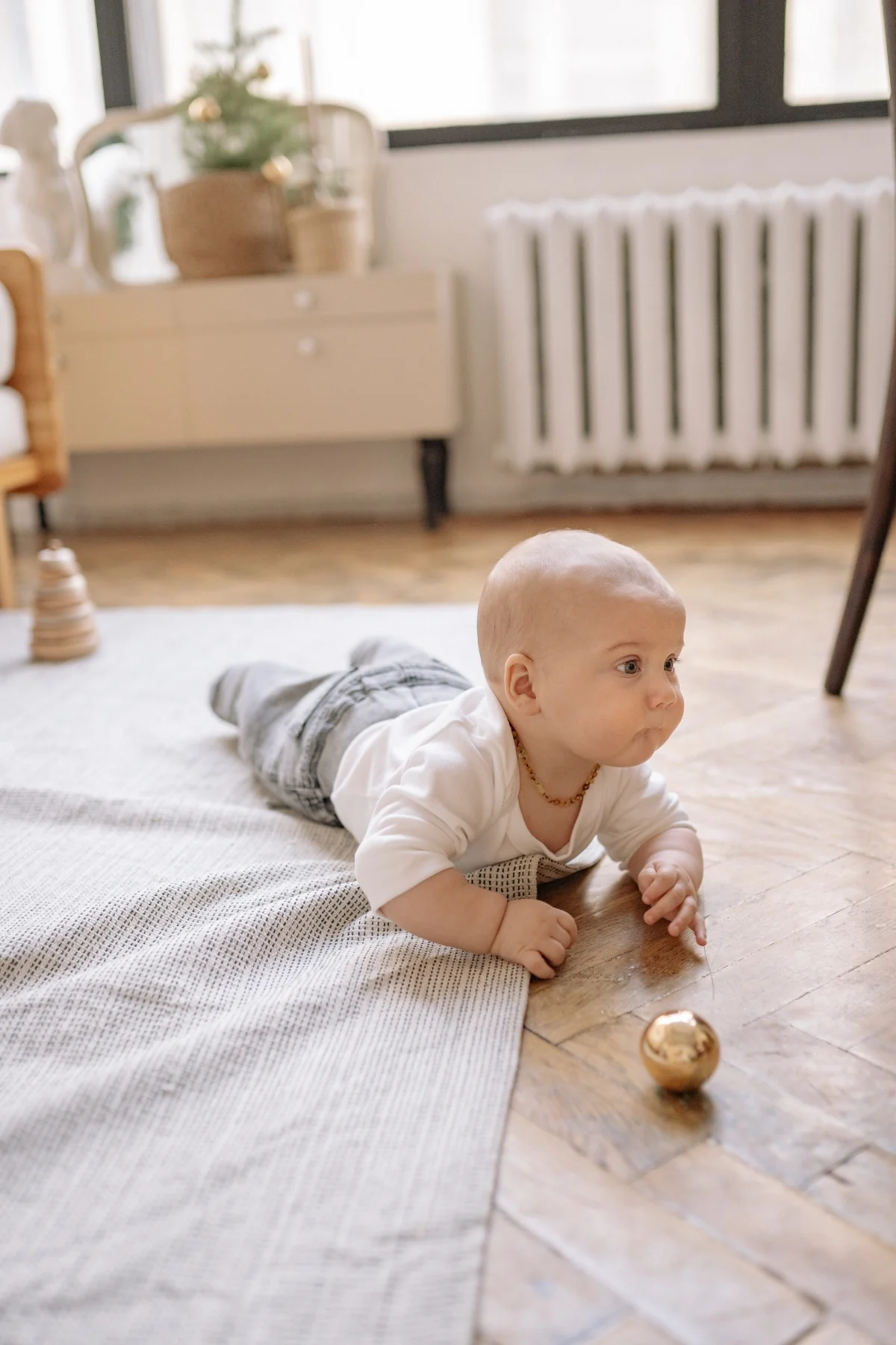 A baby lies on their tummy at the edge of a soft rug on a herringbone wooden floor in a bright room