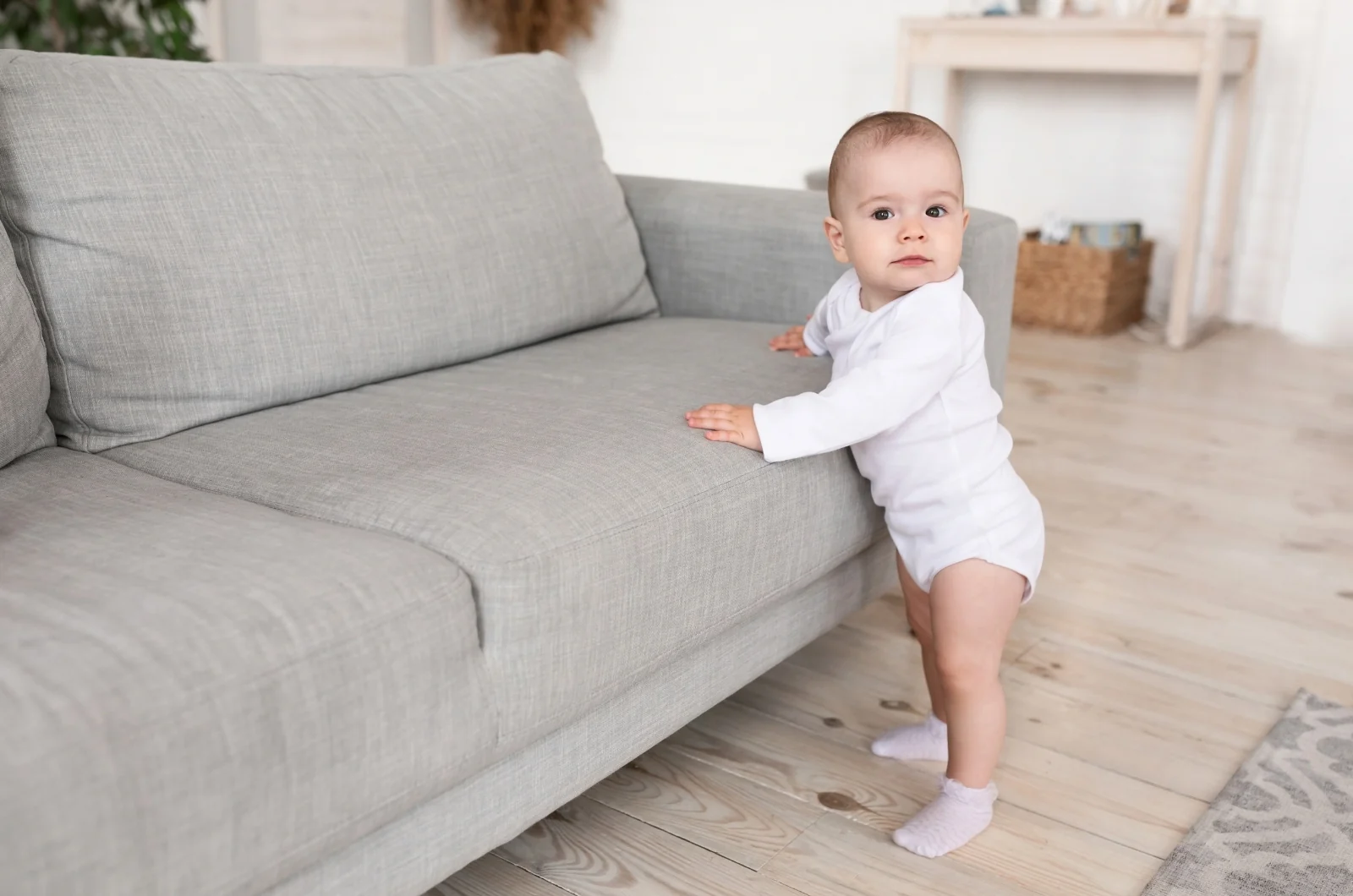 Baby standing at a gray couch, holding on with both hands