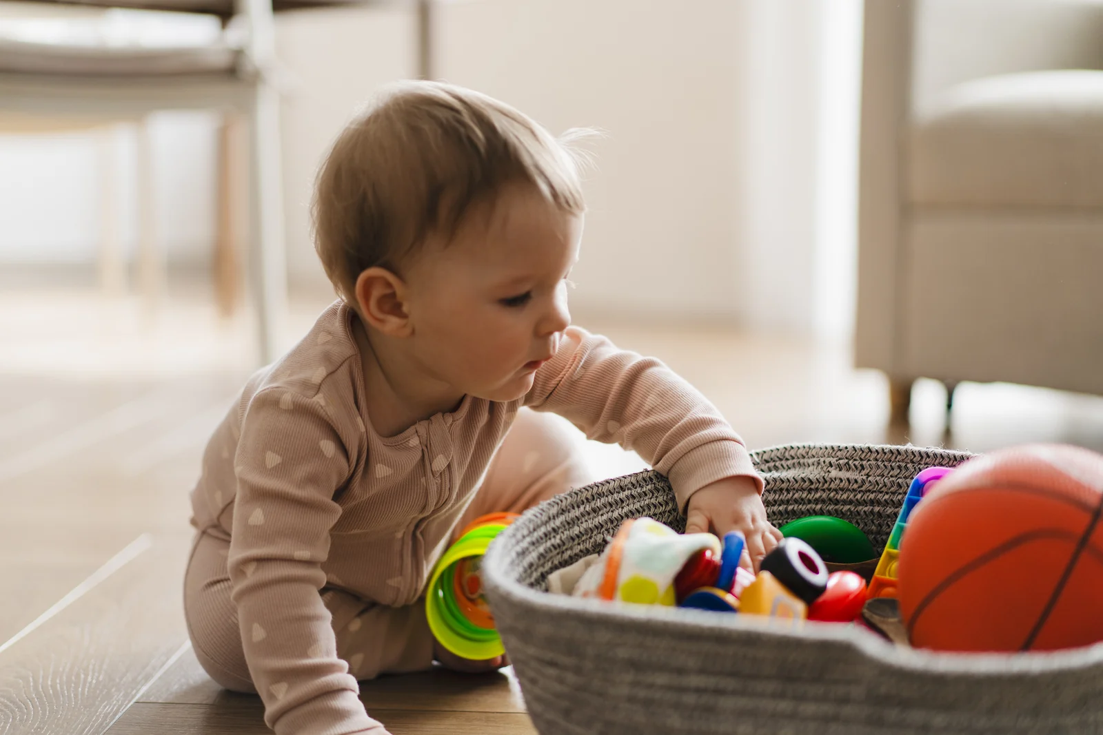 A baby sitting on a wood floor reaches into a woven basket of colorful toys