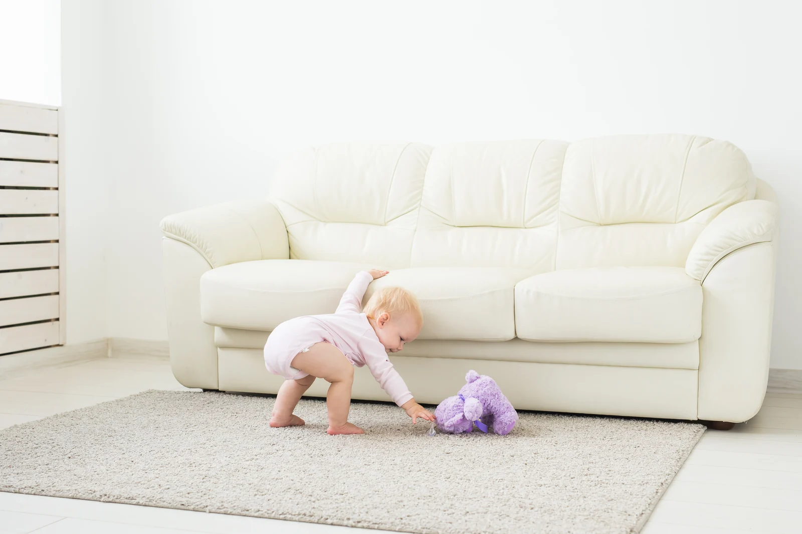 A baby in a pink bodysuit reaching for a purple plush toy on a cream rug in front of a white sofa
