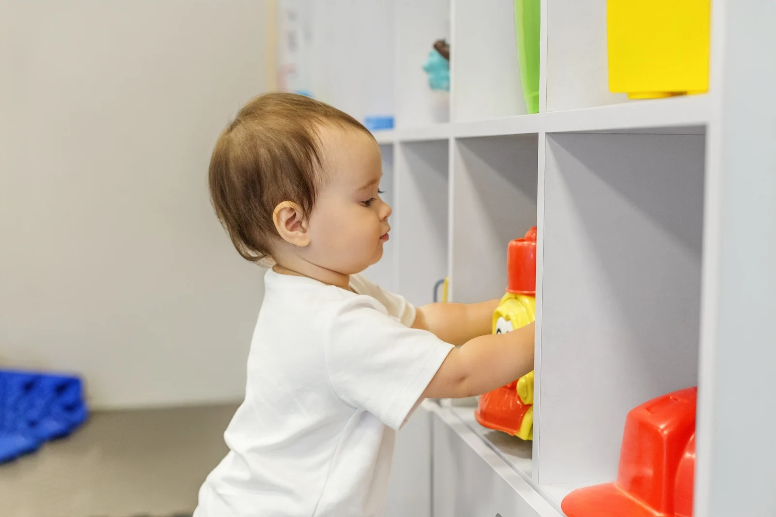 A toddler in a white tee stands at a white cube shelf and reaches for a red toy on a lower shelf