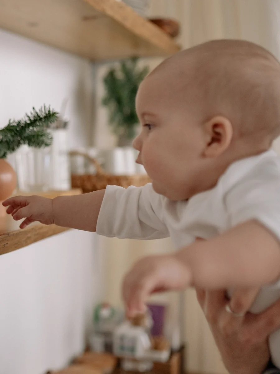 A baby held up by an adult reaches one hand toward a wooden wall shelf