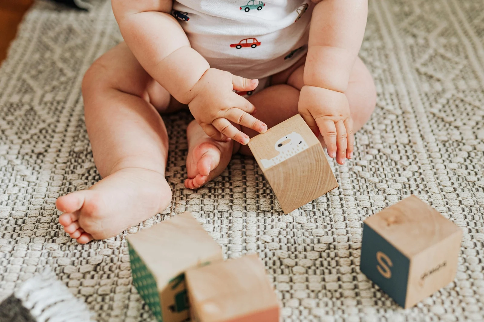A baby sits on a woven rug with one hand on a wooden block and a few more blocks nearby