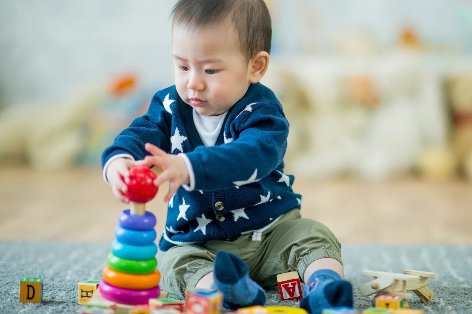 A baby in a blue star cardigan sits on a rug and places a red ring on a colorful ring stacker