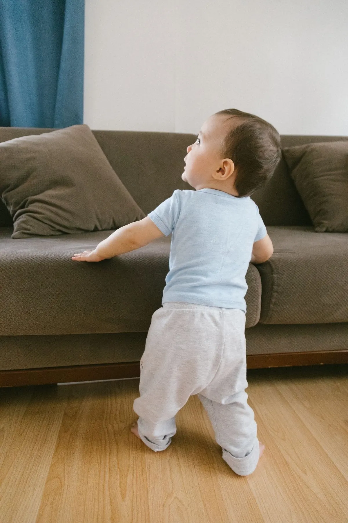 A baby in a light blue tee stands on a wood floor with one hand on the edge of a gray sofa, looking up