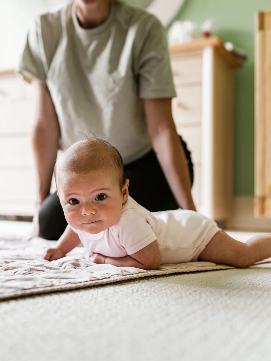 Young baby on their tummy on a rug, lifting their head, with an adult kneeling close behind