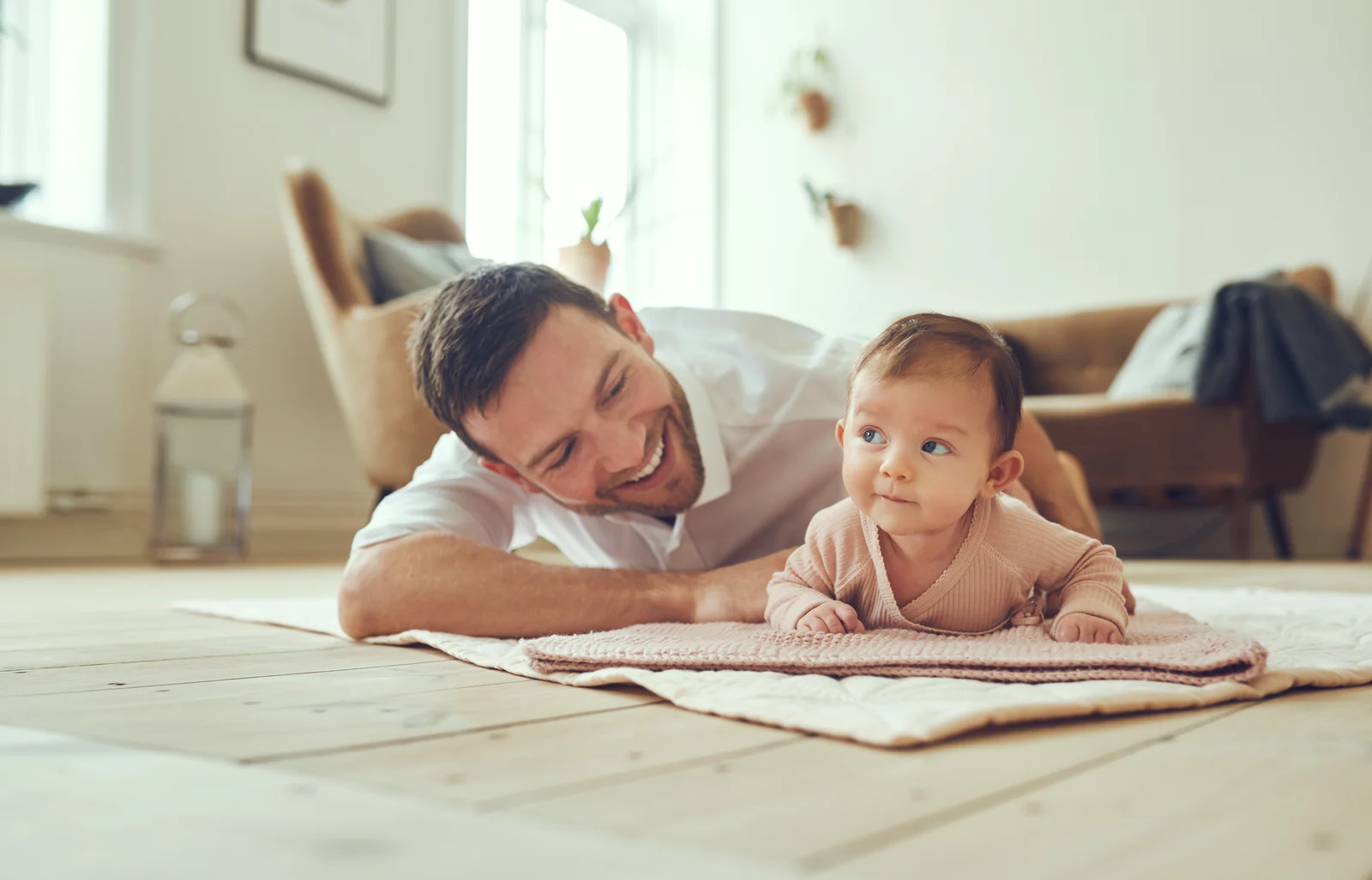 Baby on their tummy on a play mat with head up, looking aside while a smiling parent lies on the floor beside them