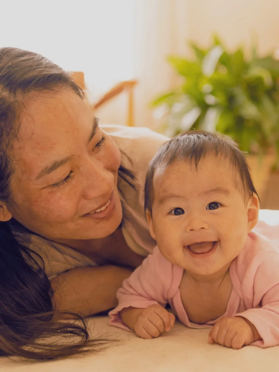 Mother lying on the floor beside a smiling baby on their tummy on a cream rug
