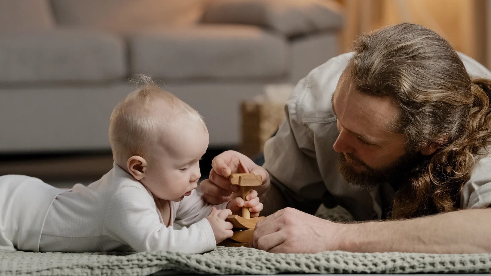 Baby lying on a knit rug, watching closely as a man's hands hold a wooden toy