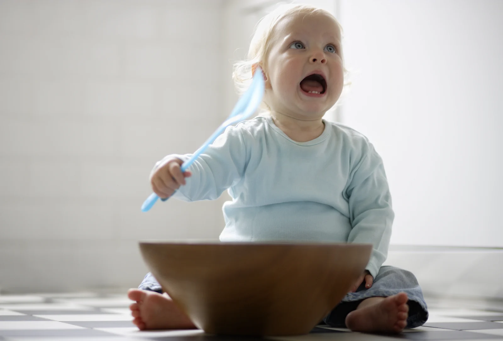 Baby sitting on a tile floor behind a large wooden bowl, waving a blue plastic spoon with a wide-open mouth