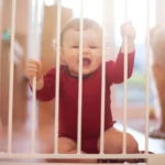 A baby in a red bodysuit sits behind a white safety gate with both hands on the bars: babyproofing