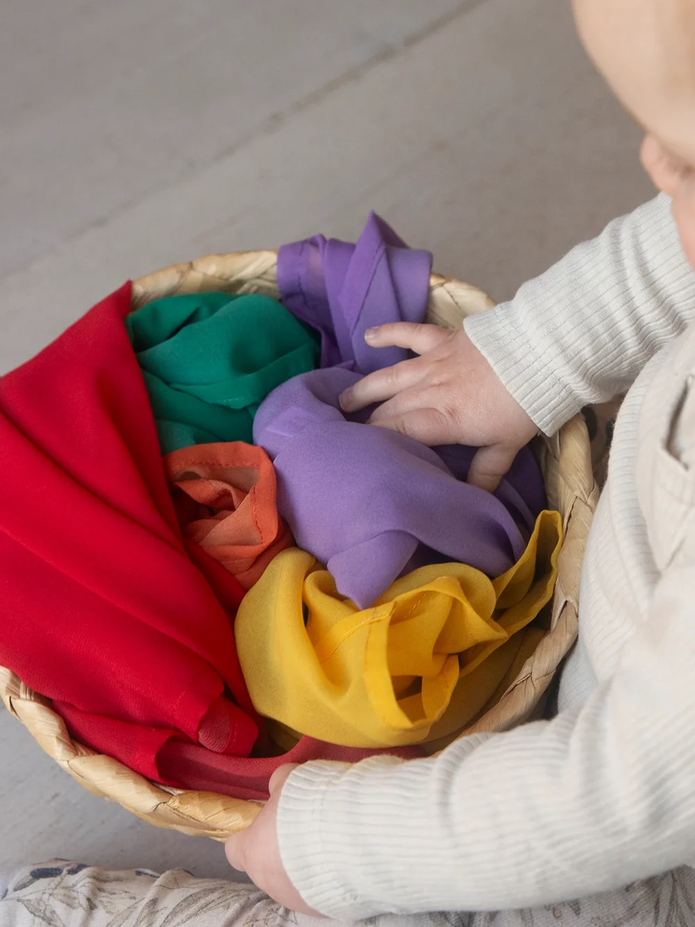 Baby sitting on the floor with a woven basket of soft play cloths in red, green, purple and yellow