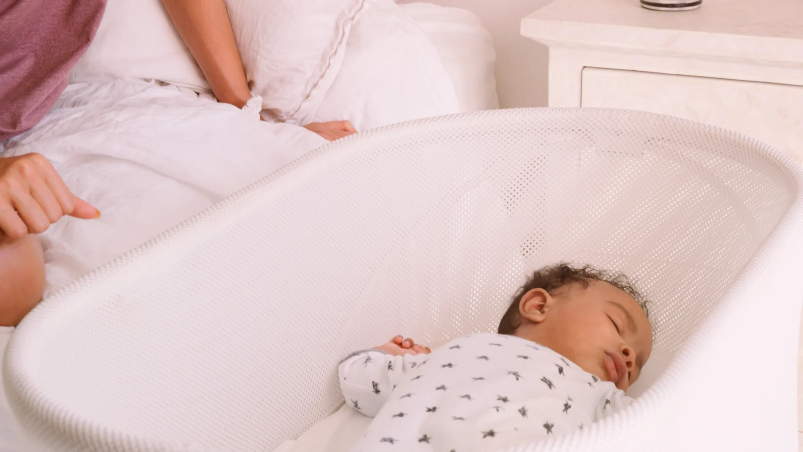 Sleeping baby in a white mesh bassinet pulled up beside a parent's bed