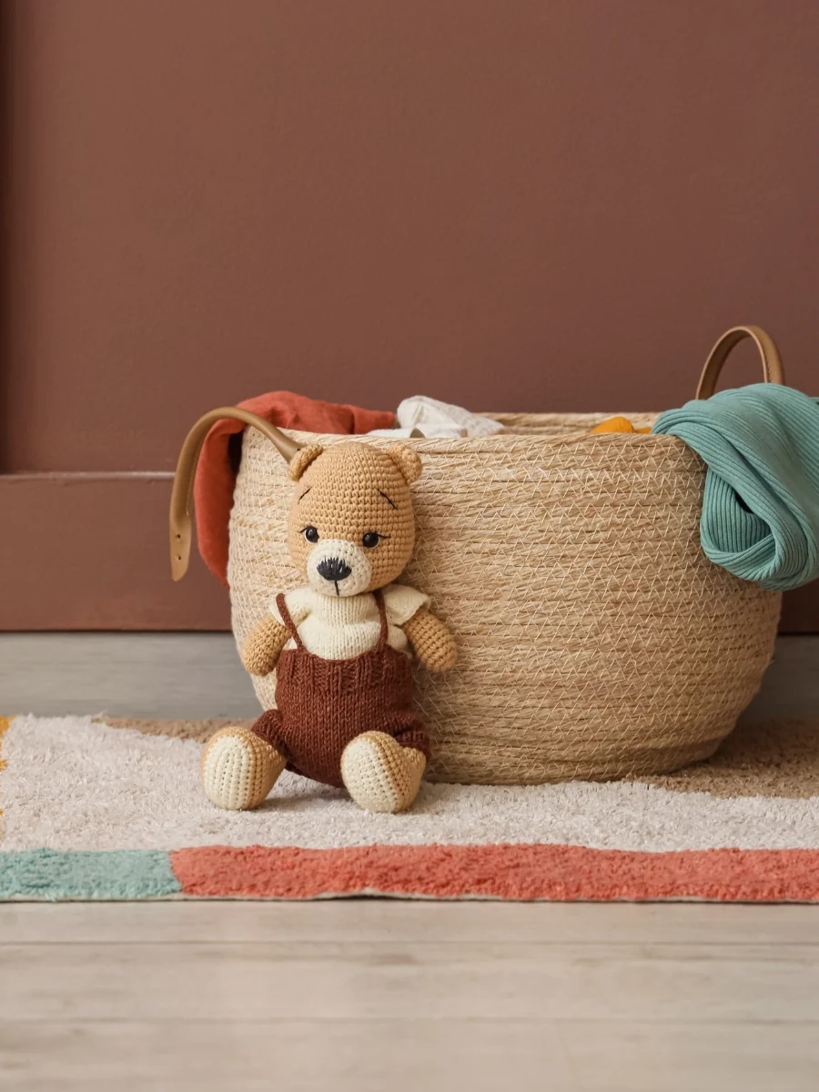 A large woven basket with folded blankets and a knitted teddy bear on a colorful rug against a brown wall
