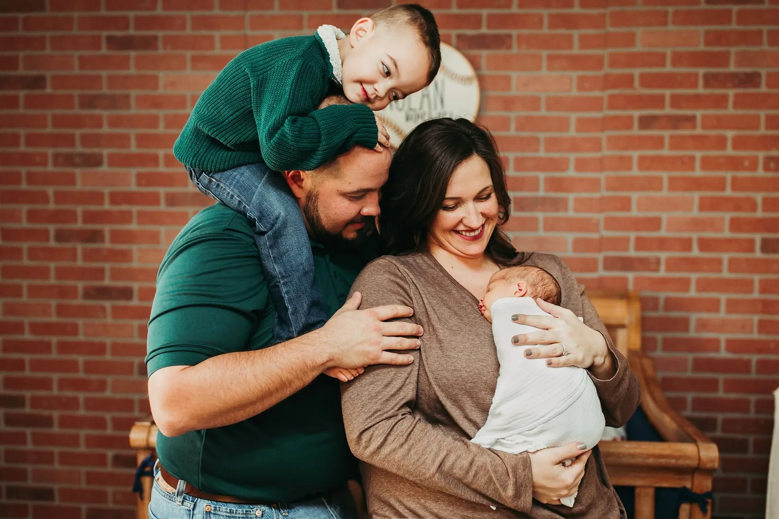 Boy riding on a man's shoulders, smiling down at a swaddled newborn in a woman's arms, in front of a brick wall