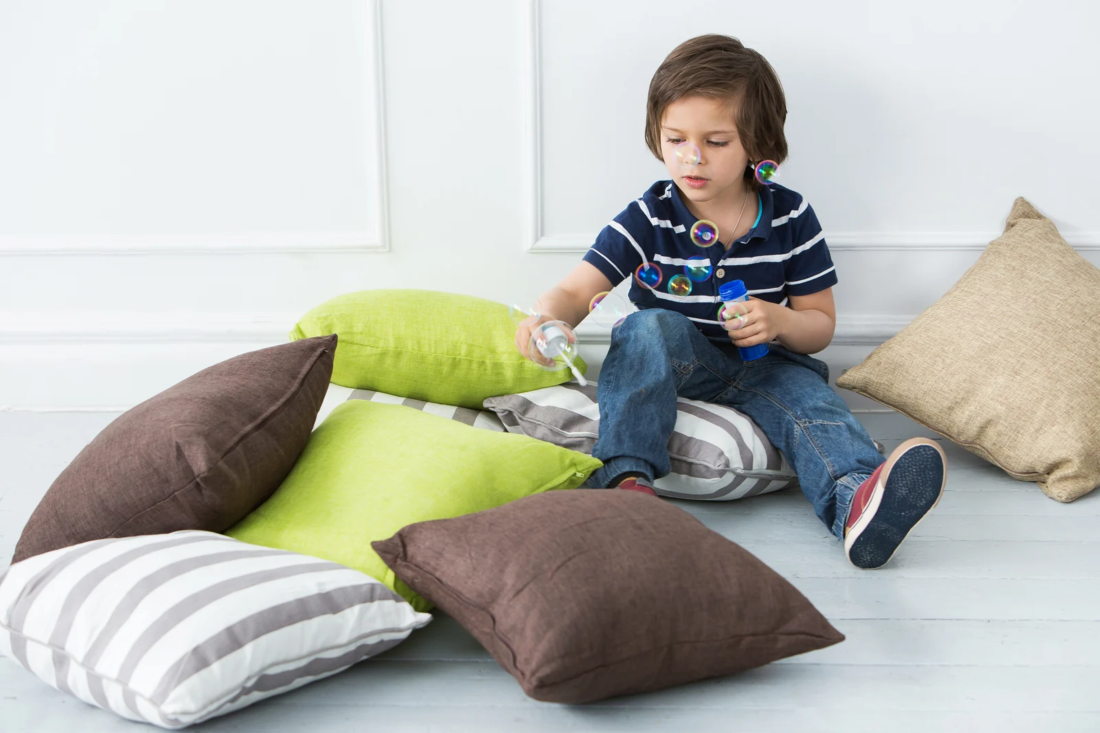 Young boy sitting on the floor blowing bubbles among scattered sofa cushions in a bright room