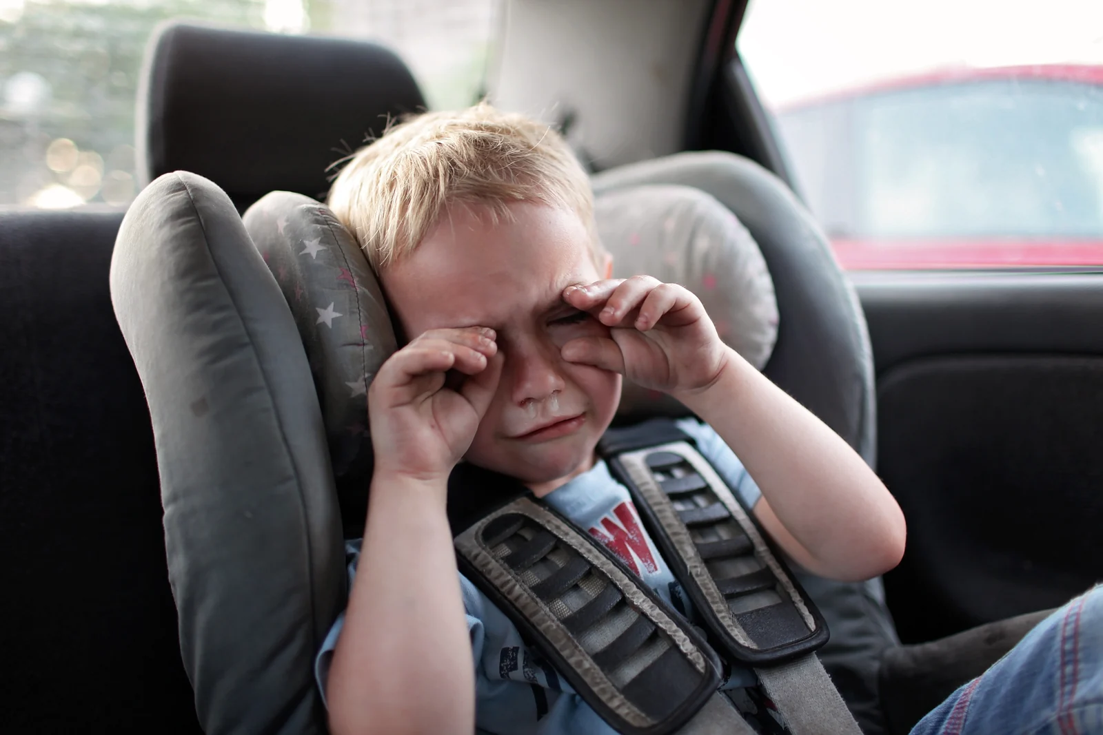 Young boy in a car seat in the back of a car, rubbing his eyes with both fists and crying
