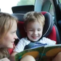 Smiling boy in a car seat looks at a picture book while a woman leans in beside him: car ride activities for kids