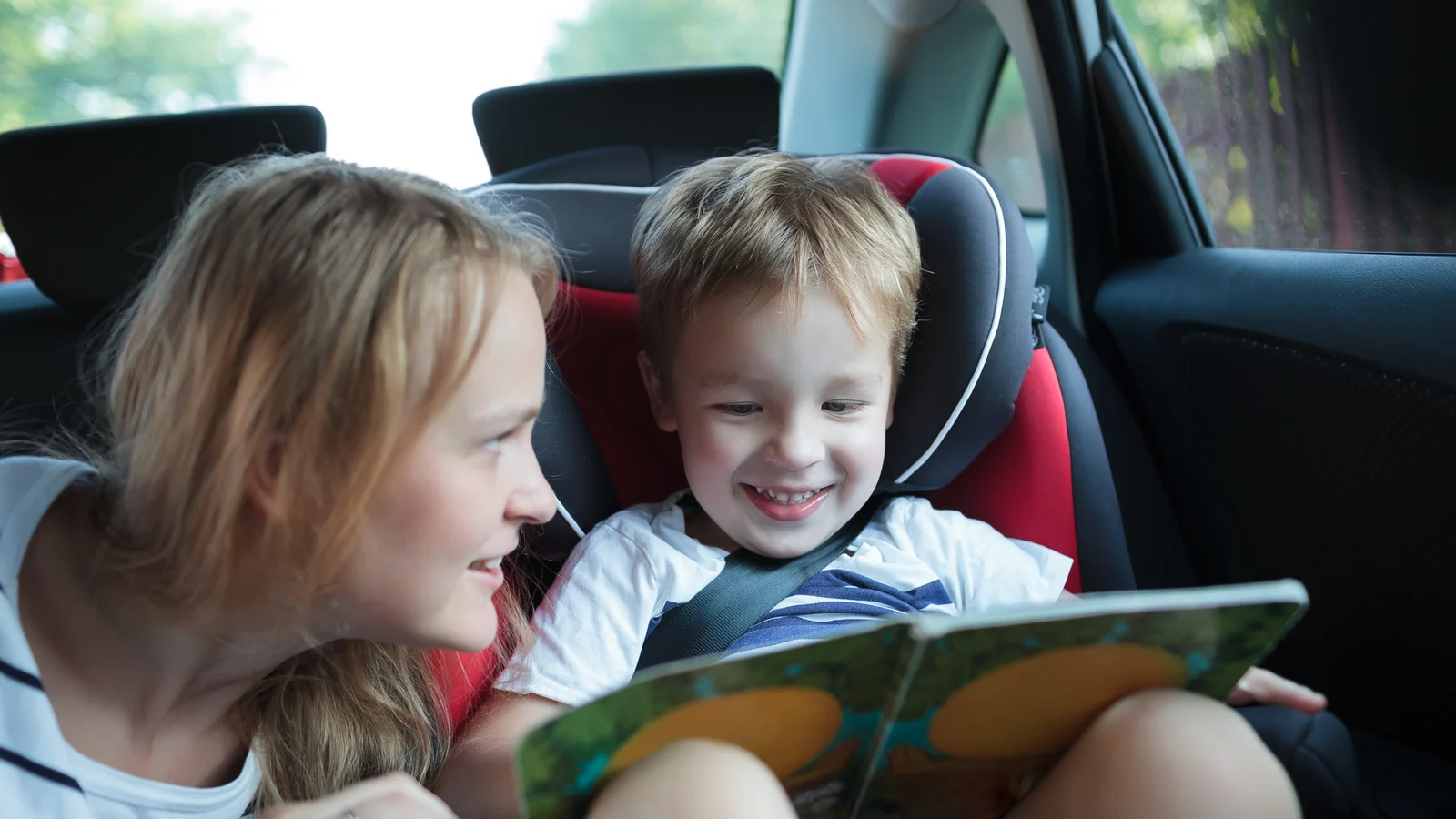 Smiling boy in a car seat looks at a picture book while a woman leans in beside him: car ride activities for kids