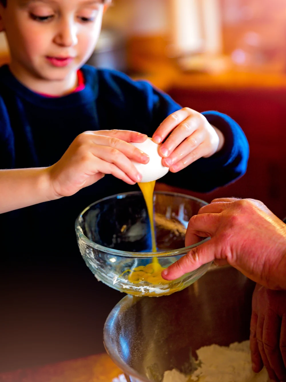 Toddler's hands cracking an egg into a mixing bowl on a low counter, with a bit of egg spilled beside it