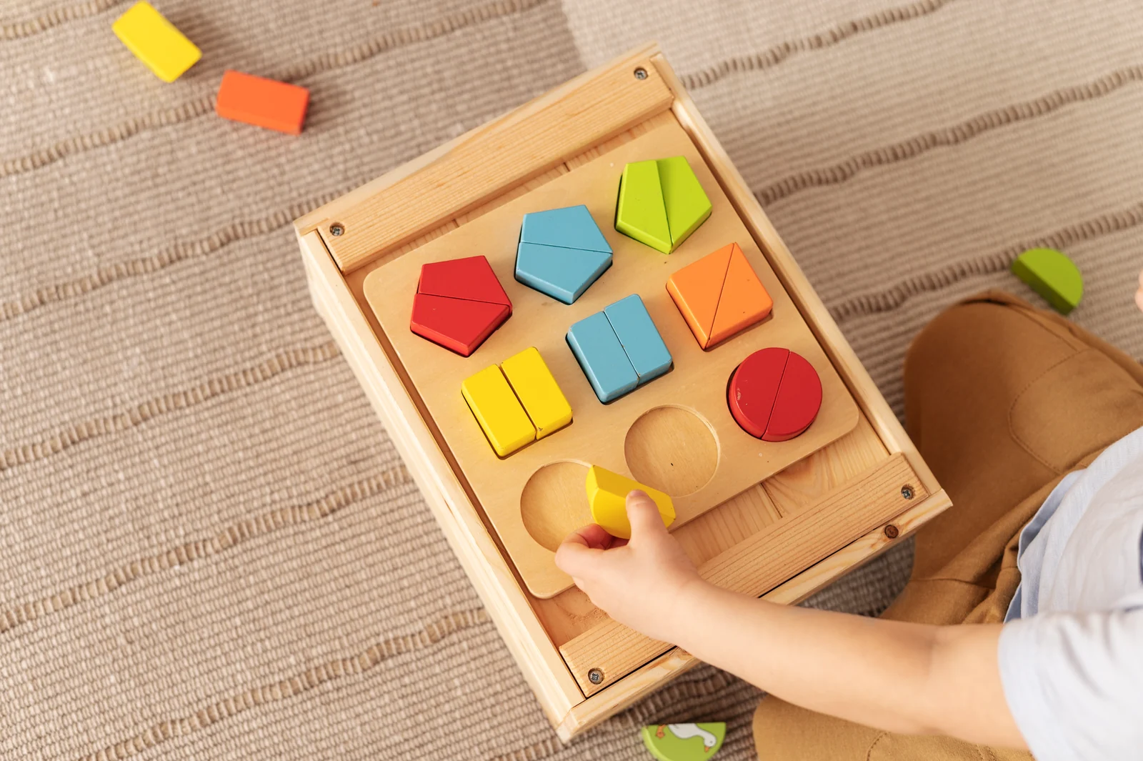 A child's hand places a yellow piece on a wooden shape sorter box with colorful shapes, on a woven rug