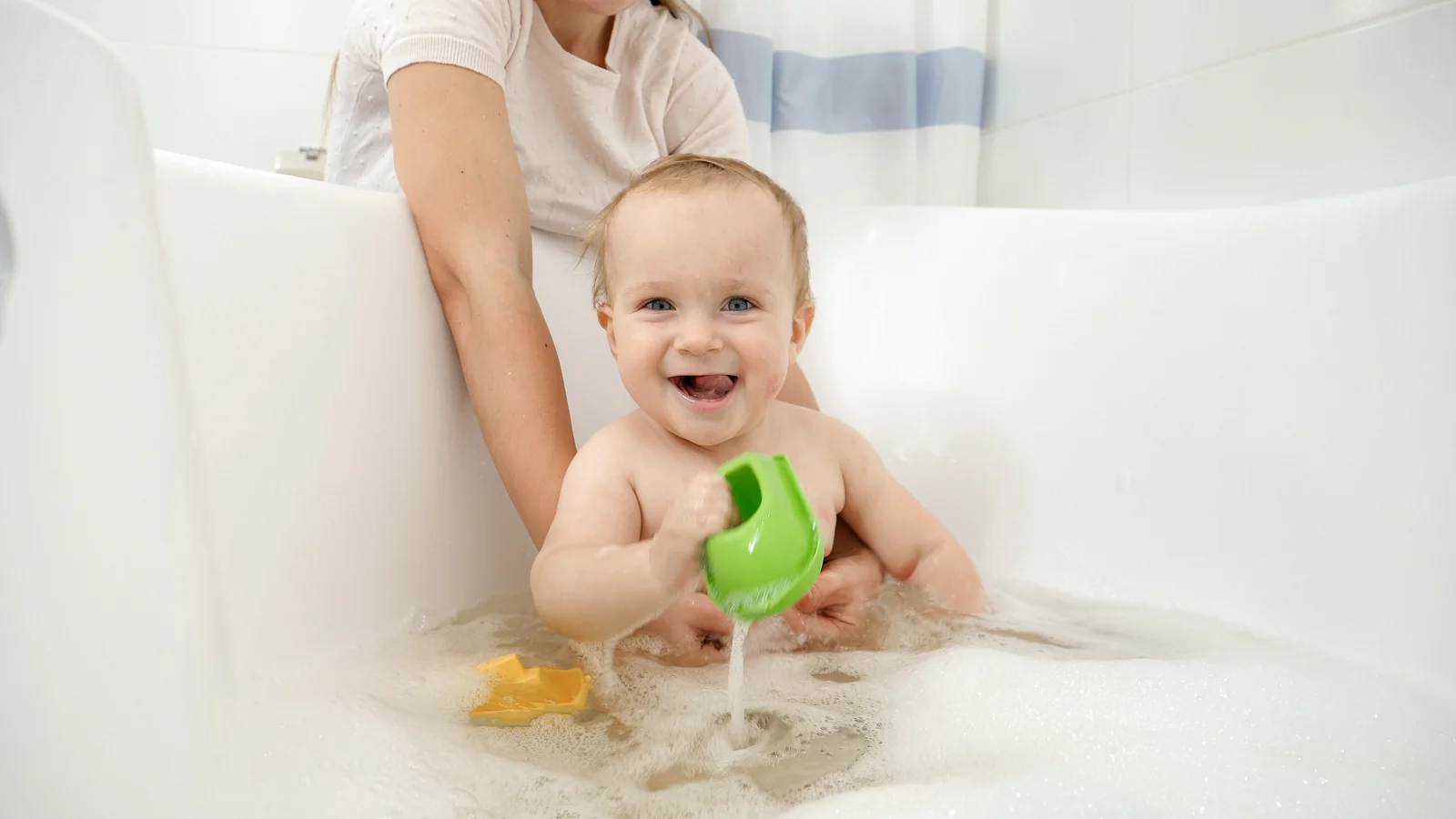 Smiling baby in a bubble bath tipping a green cup while an adult's hands steady them from behind