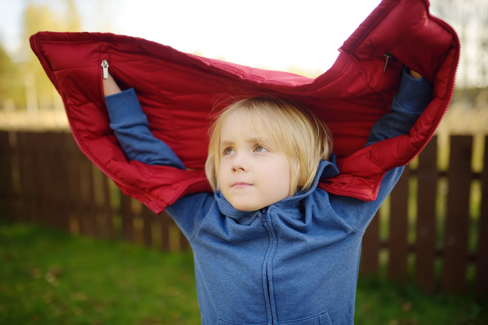 Child outdoors pulling a red puffer jacket up and off over their head, with a blue hoodie underneath