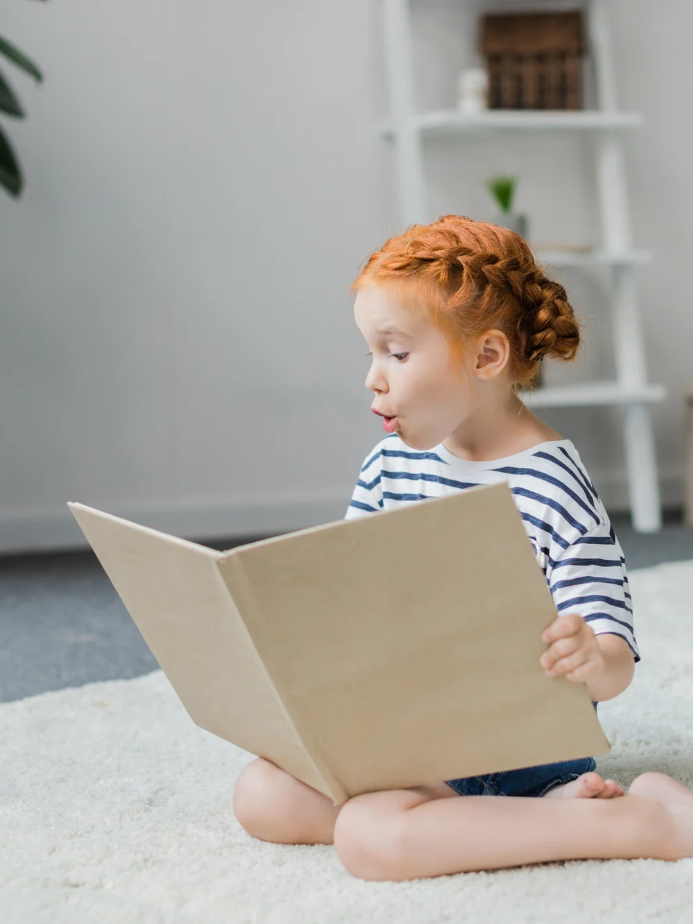 A young girl with braided red hair sits on a soft white rug holding a large open book, with a white ladder shelf behind