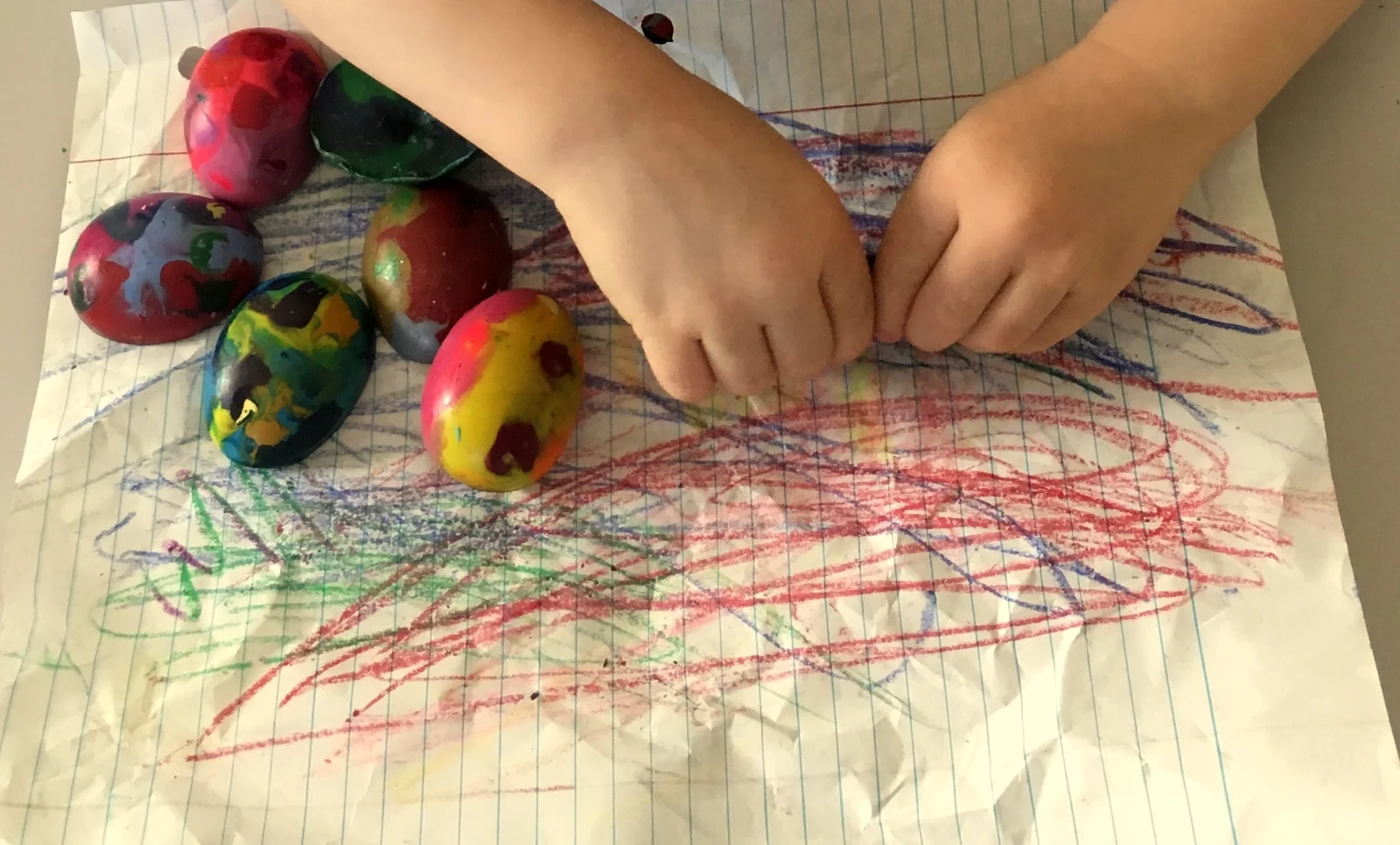 Toddler's hands on lined paper covered in crayon scribbles, with chunky, egg-shaped crayons in swirled colors beside them